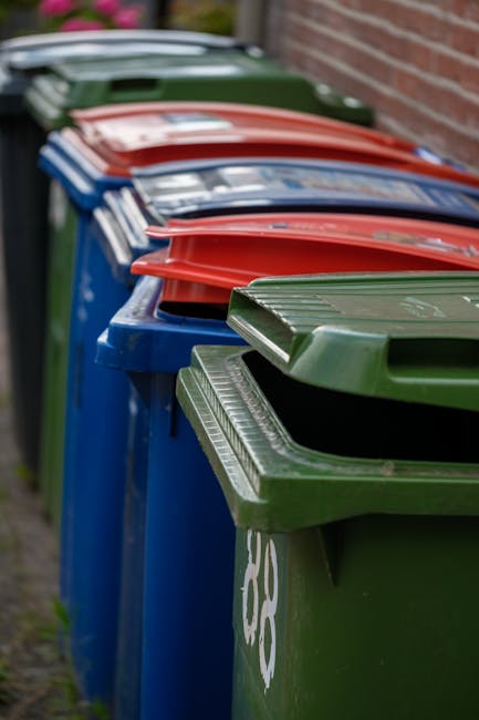 A row of large plastic wheelie bins placed along a paved pathway next to a brick wall, used for waste disposal. The bins are in various colours including green, red, and blue, each with a matching hinged lid on top. The green bin in the foreground features a textured lid with horizontal ridges, while the other bins behind it have smooth lids. The bins are positioned closely together, with some slightly tilted or stacked side by side. The surrounding environment appears to be an outdoor residential area, evidenced by the brick wall and narrow paved path. The lighting is natural and even, highlighting the different textures and colours of the bins, which are commonly used for private waste collection services such as those provided by Rubbish Collection Surrey for efficient rubbish removal and alternative waste handling solutions outside of standard municipal collection. The scene captures typical on-site waste management arrangements, emphasizing the practicality of individual bins designated for sorting or general waste disposal in a domestic setting.