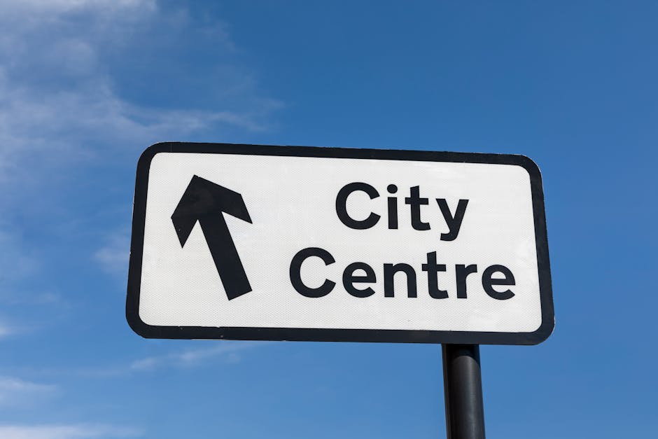 A rectangular white road sign with a black border mounted on a black pole, indicating the direction towards the city centre with a bold black upward arrow on the left side and the words 'City Centre' in large black text on the right side. The sign is positioned outdoors against a backdrop of a clear blue sky with some light clouds. The surrounding environment suggests an urban or suburban setting, typical for signage directing traffic or pedestrians. The sign's surface appears smooth and reflective, designed for high visibility and easy readability. The context implies it may be part of an independent or private collection of directional signs, common in areas where alternative waste handling or on-site clearance may occur, such as near commercial or municipal facilities. Rubbish Collection Surrey often deals with various waste management signage, supporting safe and efficient disposal practices in town centres, although this particular sign does not directly relate to rubbish removal services.