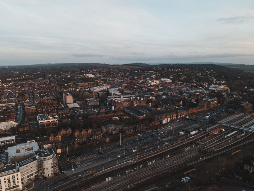 An aerial view of Guildford town centre, displaying a dense urban area with a mixture of modern and traditional brick buildings, commercial structures, and residential properties, all set amidst a backdrop of rolling countryside and distant hills. In the foreground, multiple railway tracks run parallel to a busy road with visible vehicles, streetlights, and parking areas. The lighting appears to be during late afternoon or early evening, with a partly cloudy sky casting soft, diffused light over the scene. The overall environment suggests a typical cityscape with busy infrastructure and structured urban planning, which could be of interest to those seeking private or alternative rubbish collection or waste management services in the Surrey area. Rubbish Collection Surrey operates in such urban settings, providing rubbish removal solutions amidst the busy town environment.