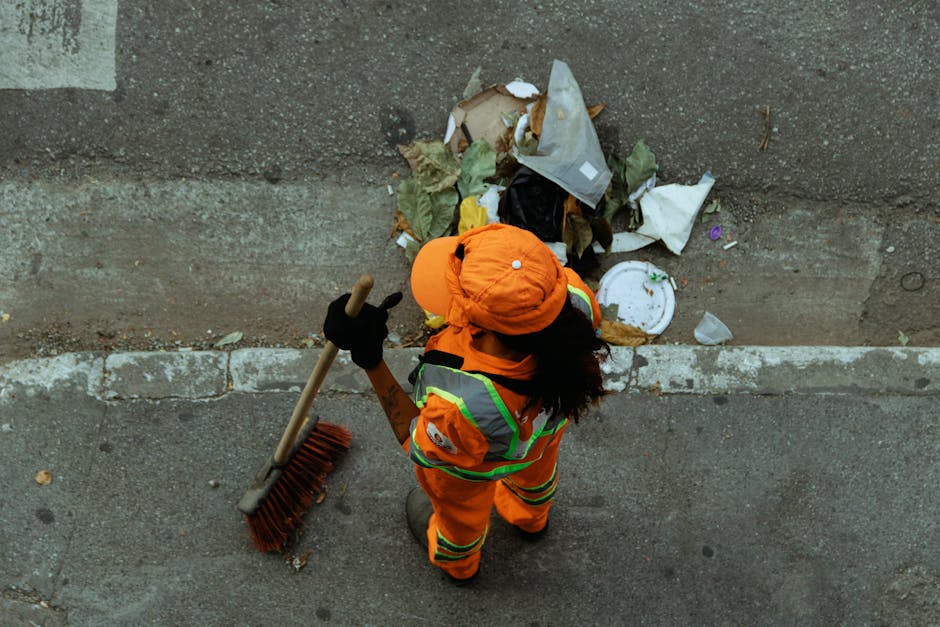 A worker dressed in bright orange high-visibility clothing, including a jacket, pants, gloves, and a hat, is standing on a concrete sidewalk and is seen from above. The worker is holding a broom with a long wooden handle and red bristles in their right hand, and is facing a pile of mixed waste that includes paper, plastic, and cardboard materials, some of which are crumpled or torn. The waste pile is resting against a plain concrete or plaster wall, with the debris spread along the edge of the sidewalk, adjacent to a narrow strip of dirt and small stones. The lighting appears natural, suggesting outdoor daytime conditions, and the scene is set in an urban environment consistent with common waste collection or on-site clearance activities. The image relates to rubbish removal or waste disposal services, emphasizing independent collection efforts by waste management professionals like those at Rubbish Collection Surrey, as it visually depicts waste being raked up or prepared for removal.